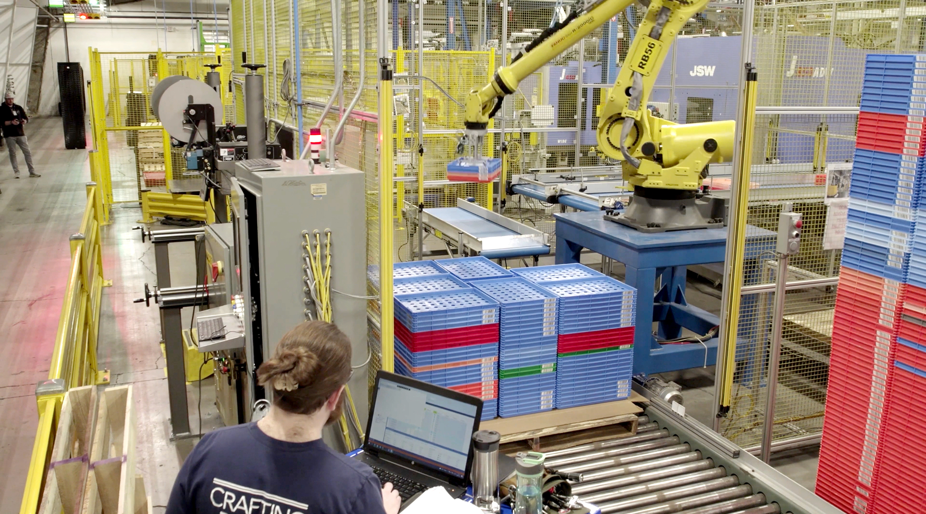 Technician monitors robotic arm stacking plastic trays on a conveyor inside an automated manufacturing facility.