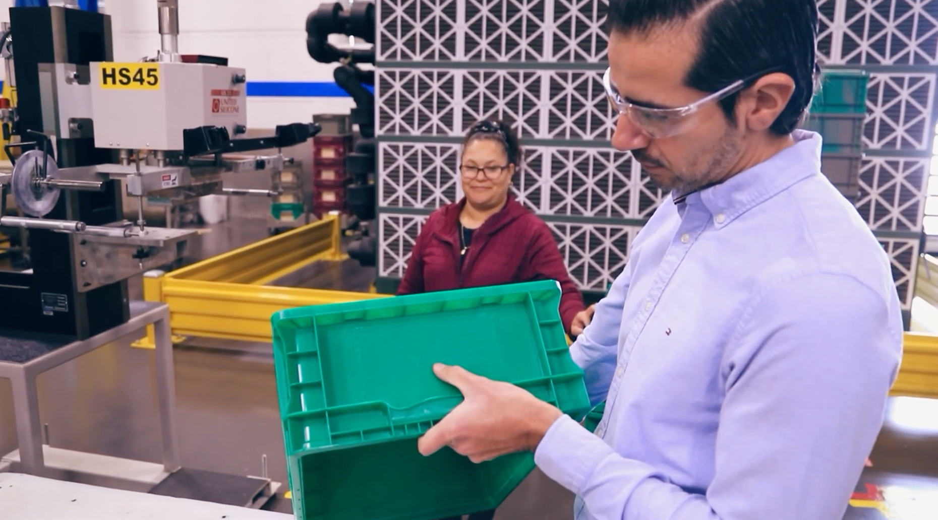 Engineer inspecting a green plastic container while a coworker observes in a manufacturing facility.