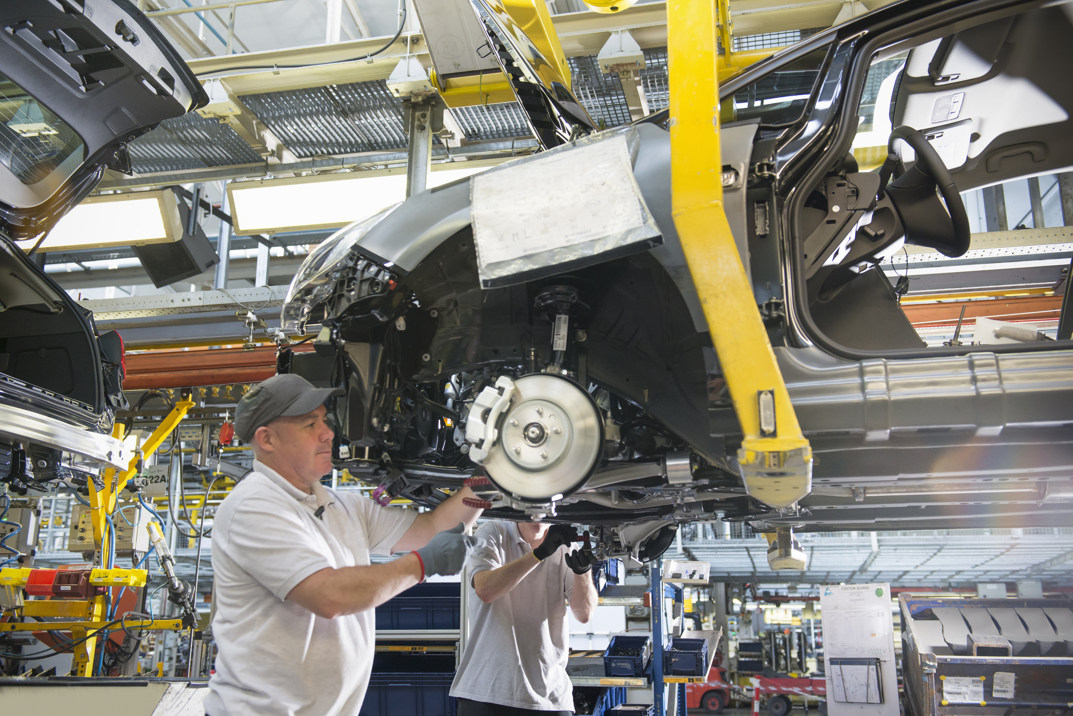Workers assemble a car chassis on an automotive production line inside a factory with overhead lifting equipment.