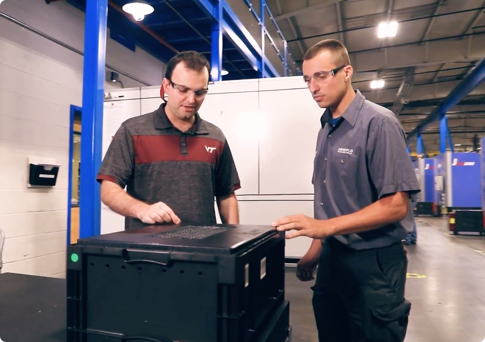 Two workers in safety glasses inspecting a black plastic tote inside a manufacturing facility.