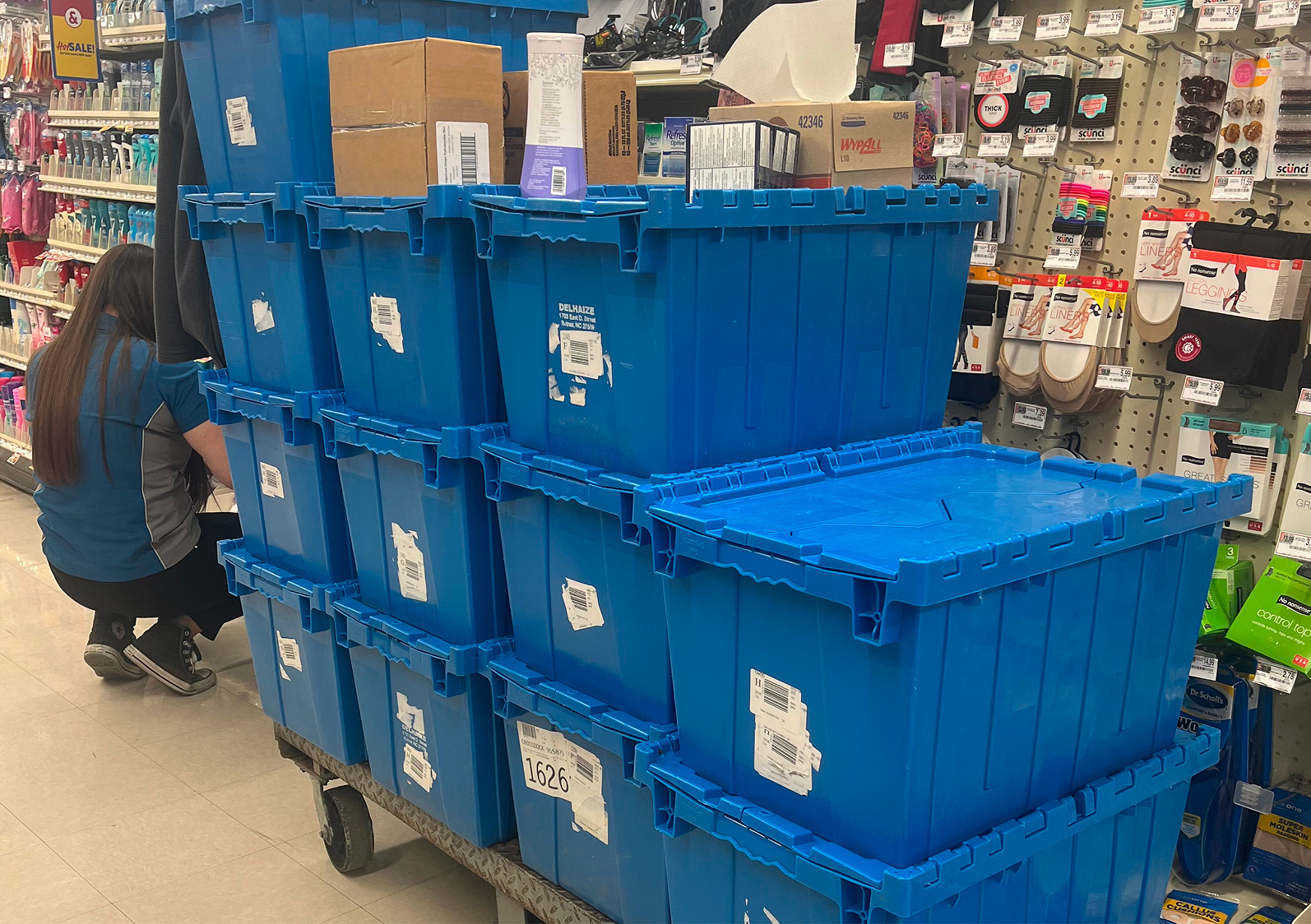 Store employee stocking shelves beside stacked blue plastic totes on a cart in a retail aisle.